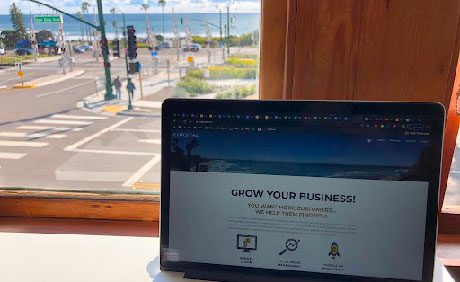 A laptop displaying a business website is positioned on a desk near a window, with a clear view of a street and the ocean in the background.
