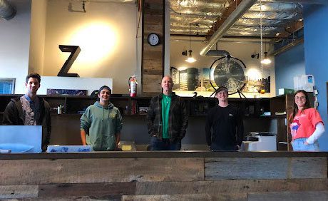 Five people stand behind a wooden counter in a room with industrial decor, including exposed ducts and a clock.