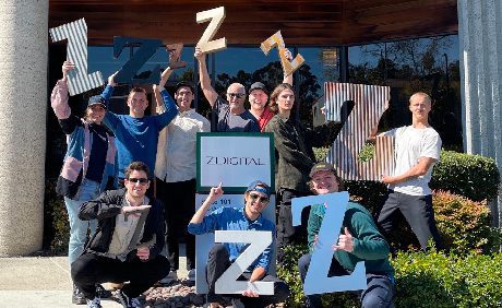 A group of people standing outside a building, holding large letter Z cutouts. A sign reading, ZDIGITAL, is displayed in the center.