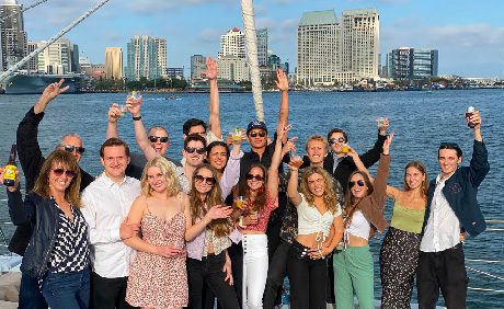 Group of people smiling, holding drinks, and posing with arms raised on a boat, with a city skyline and waterfront in the background.