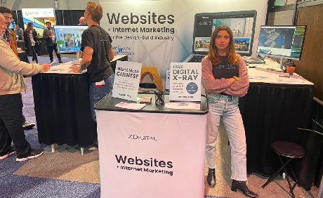 A woman stands at a booth labeled, Websites + Internet Marketing, with promotional signs for services at a trade show. People are interacting in the background.