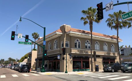 Street corner with a beige, two-story building featuring arched windows. Traffic lights and street signs are visible, with cars parked along the road. Palm trees line the sidewalk.