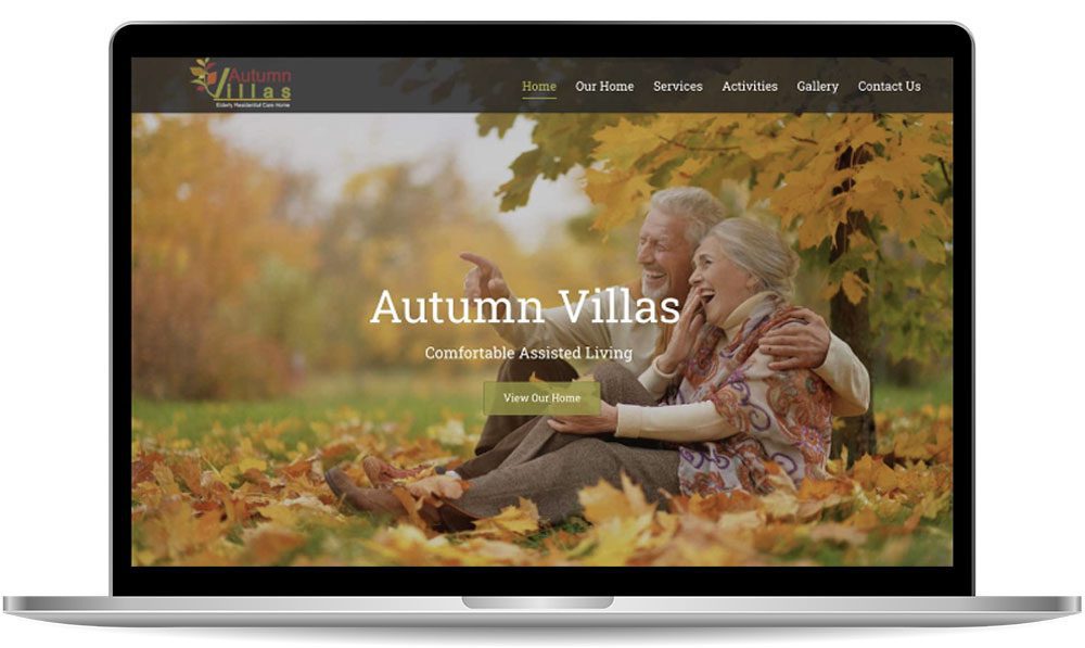Laptop screen displaying a webpage for Autumn Villas, showcasing an image of an elderly couple sitting on grass amid fall foliage. The website's menu and a button that reads "View Our Home" are visible.
