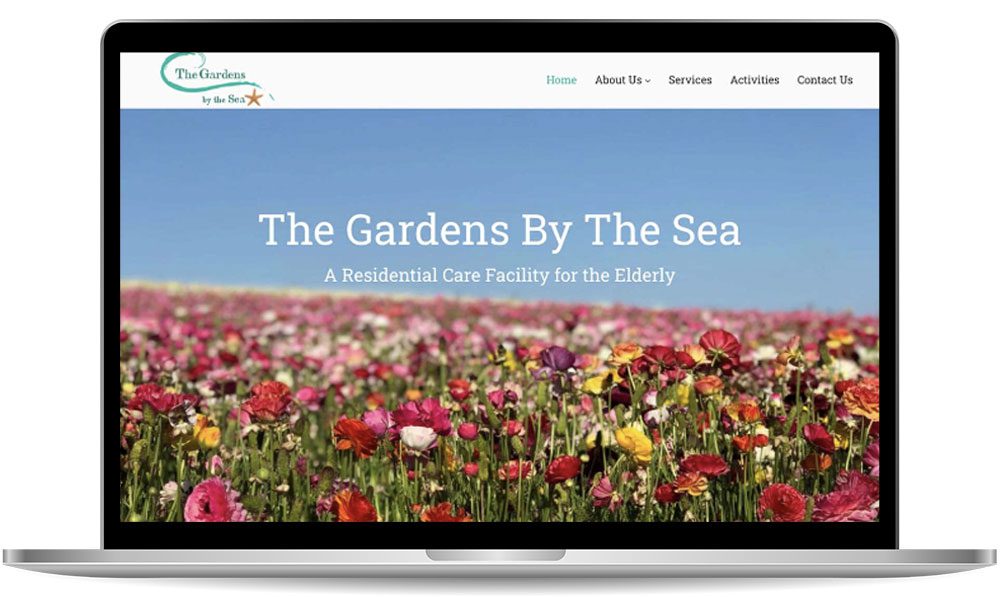 A laptop screen displays a website for "The Gardens By The Sea," a residential care facility for the elderly, with a header image of a field of colorful flowers under a blue sky.