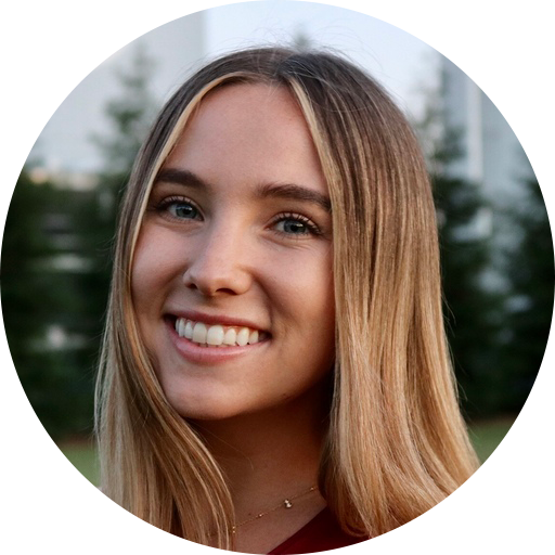 Woman with long blonde hair smiling, outdoors with trees and a building in the background.