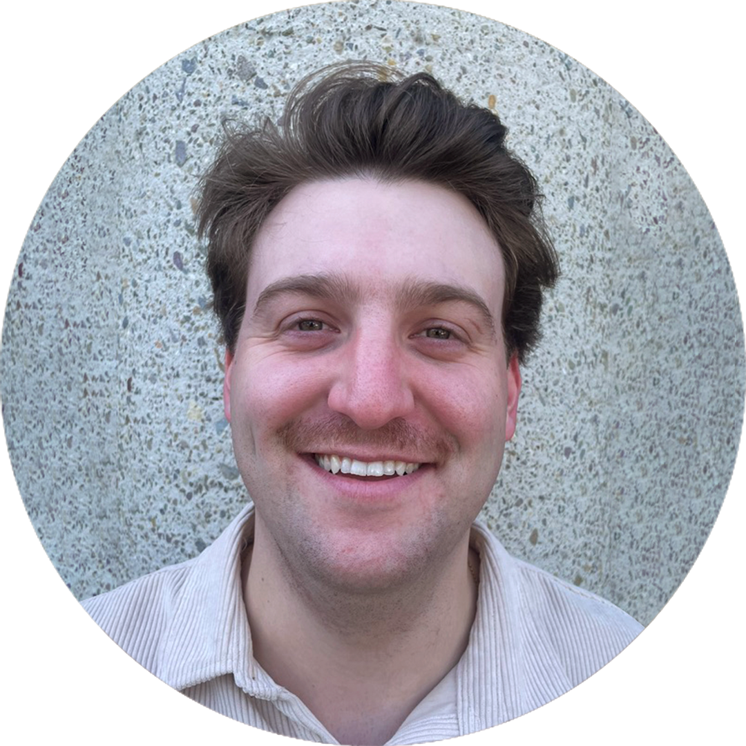 Person with a mustache smiling at the camera, standing against a textured stone wall.