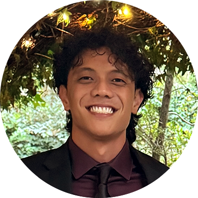 A person in a dark suit and tie smiles at the camera while standing under foliage with string lights.