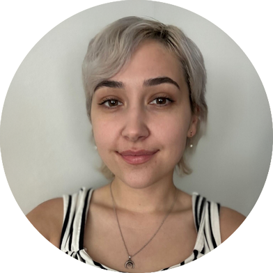 A person with short, light blonde hair and neutral expression looks at the camera. They are wearing a striped top and a necklace with a circular pendant.