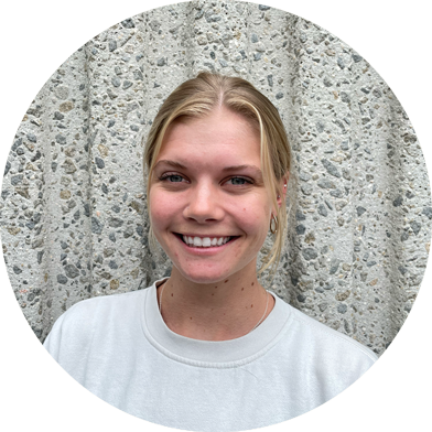 A young woman with blonde hair, wearing a white shirt, smiles at the camera while standing in front of a textured concrete wall.