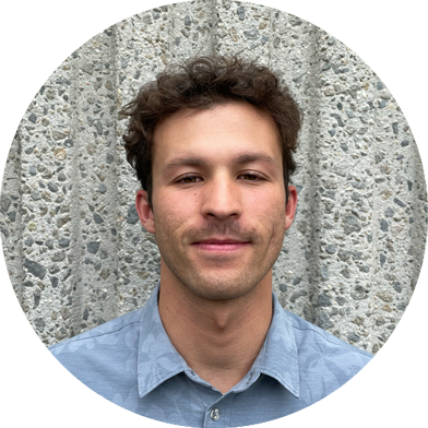 Young man with short curly brown hair and a mustache, wearing a light blue collared shirt, standing in front of a textured concrete wall.