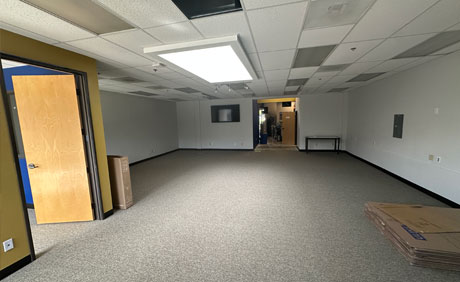 Empty office space with gray carpet, white ceiling tiles, fluorescent lighting, and some cardboard boxes near an open wooden door. A table is against the far wall.