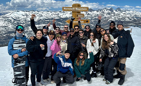 A group of people pose for a photo in the snow with snowboards and ski gear, with mountain scenery and a Mammoth Mountain sign in the background.