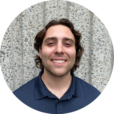 A man with wavy brown hair, wearing a dark blue collared shirt, smiles in front of a textured concrete wall.